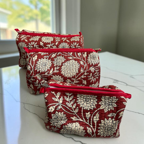 Three small zippered cosmetic pouches with a red and cream floral damask pattern, arranged on a white marble countertop near a window.