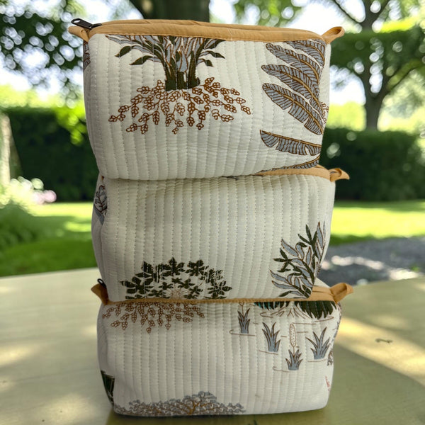 Three stacked quilted fabric zippered pouches with a botanical print, set on a wooden table outdoors with a blurred garden background.