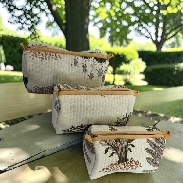 Three quilted cosmetic pouches with a brown botanical leaf pattern and tan zipper trim, arranged on a wooden bench outdoors with a blurred garden background.