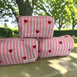 Three pink and white striped fabric pouches with red heart patterns are stacked on a wooden bench outdoors with a blurred garden background.