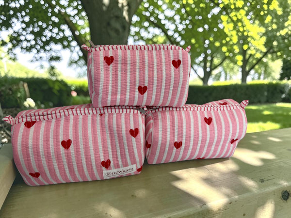 Three pink and white striped fabric pouches with red heart patterns are stacked on a wooden bench outdoors with a blurred garden background.