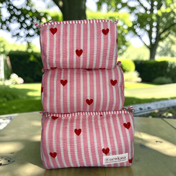 Three stacked zippered pouches in pink and white vertical stripes with small red hearts, displayed outdoors on a wooden table with a blurred garden background. A small white label on the bottom pouch reads 'newkasa'.