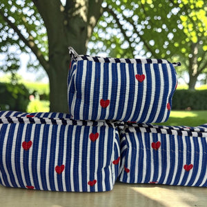 Three zippered pouches with a blue and white striped pattern and small red hearts, stacked on a light-colored wooden table outdoors with a blurred park background.