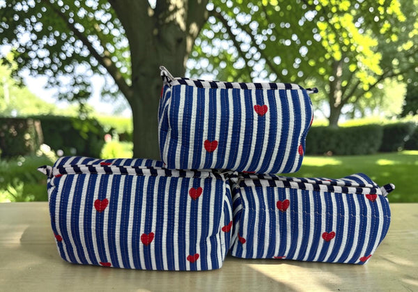 Three zippered pouches with a blue and white striped pattern and small red hearts, stacked on a light-colored wooden table outdoors with a blurred park background.