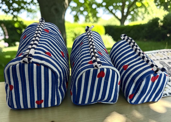 Three blue and white striped fabric pouches with red heart patterns and black-and-white braided zipper pulls, arranged on a light wooden table outdoors with trees in the background.