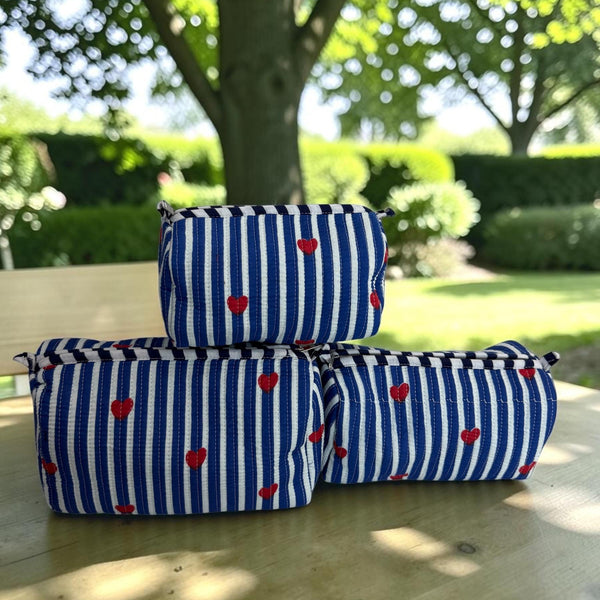 Three zippered cosmetic pouches with a blue and white striped pattern and small red hearts, stacked on a wooden table outdoors with a garden background.